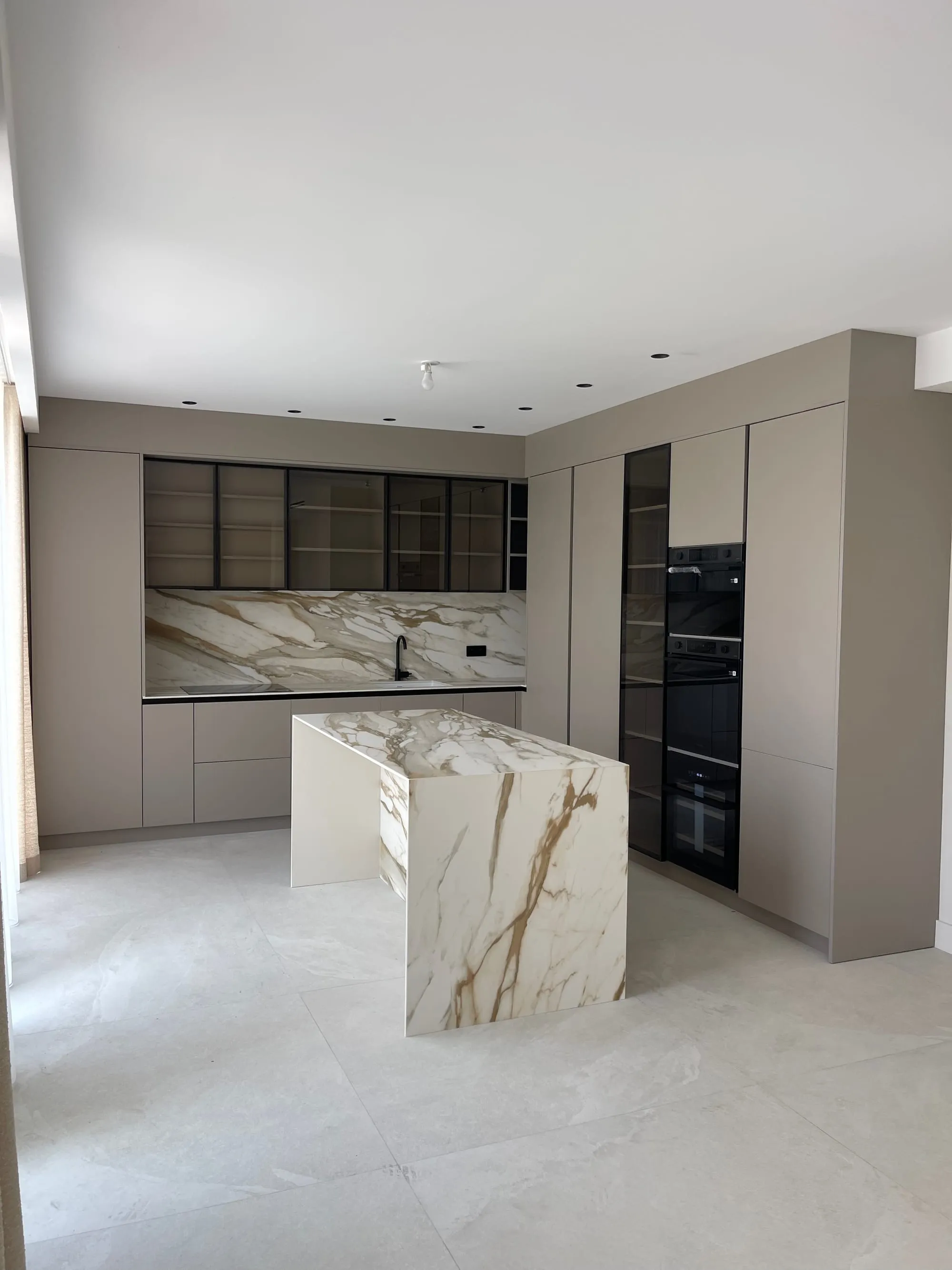 Cuisine aux façades taupe, vitrines à cadre noir, fours en colonne et îlot veiné