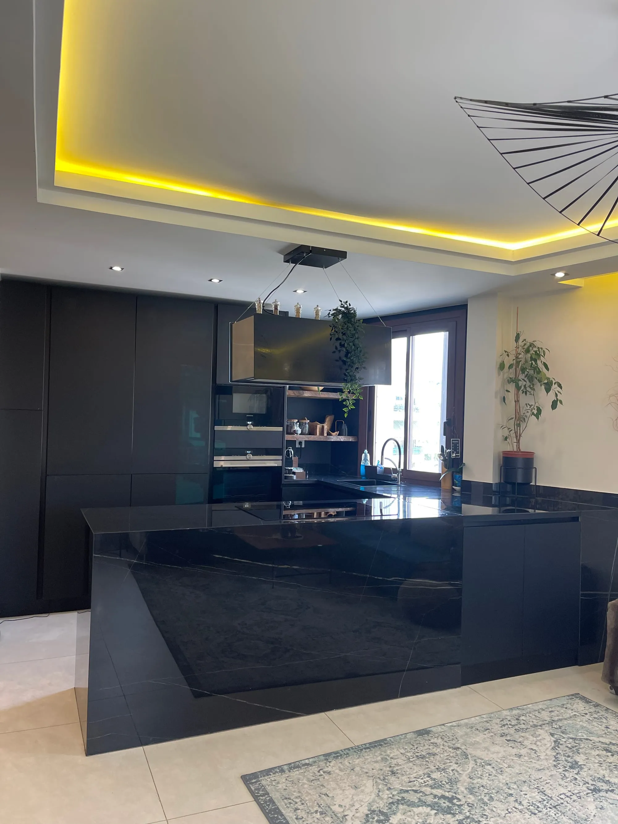 Cuisine noire avec îlot au plan brillant veiné, sous un faux plafond éclairé